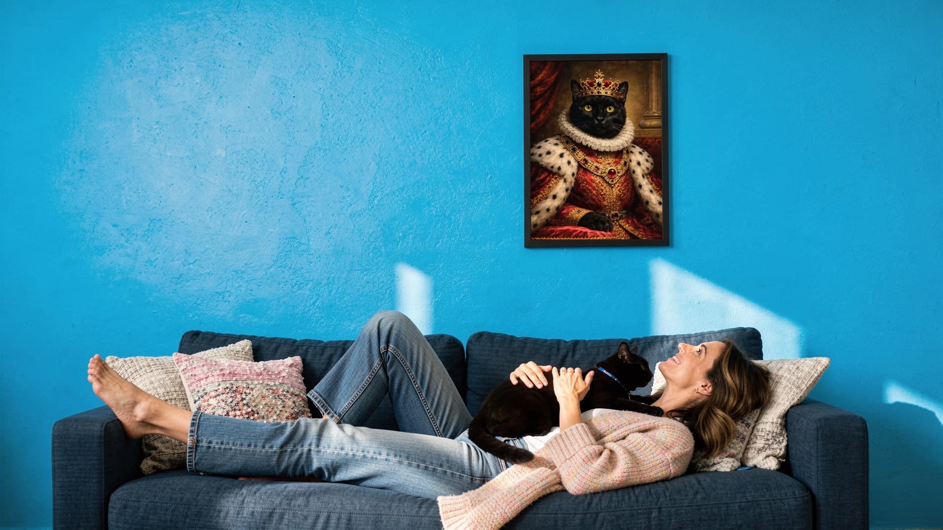 A pet parent relaxing on a sofa with her black cat, beneath a framed renaissance-style royal portrait of that same cat